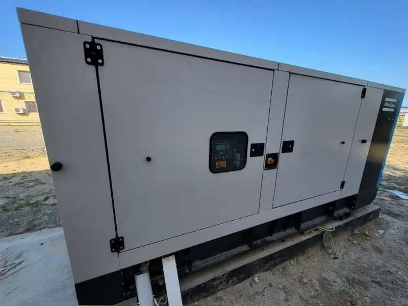 Generator von Atlas Copco in einem schallgedämmten Gehäuse mit Sichtfenster zur Steuerung in der Tür, aufgestellt auf einem Freigelände neben einem Gebäude.
